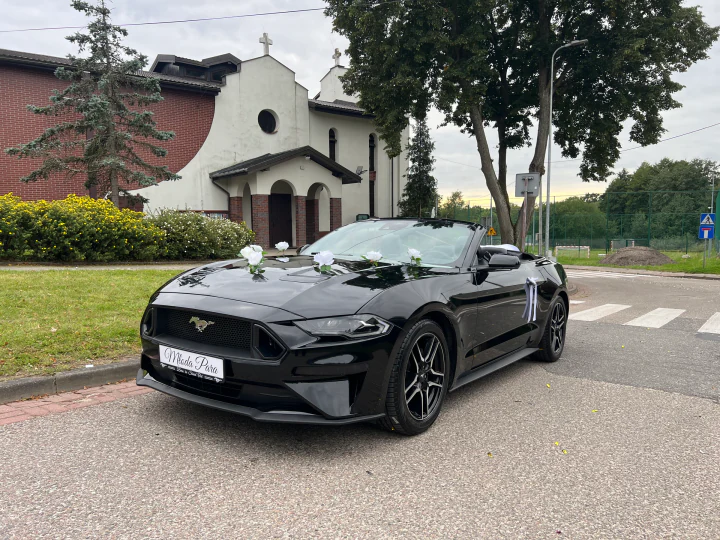 Mustang z tablicą Młoda Para i białymi kwiatami przed miejscem ceremonii